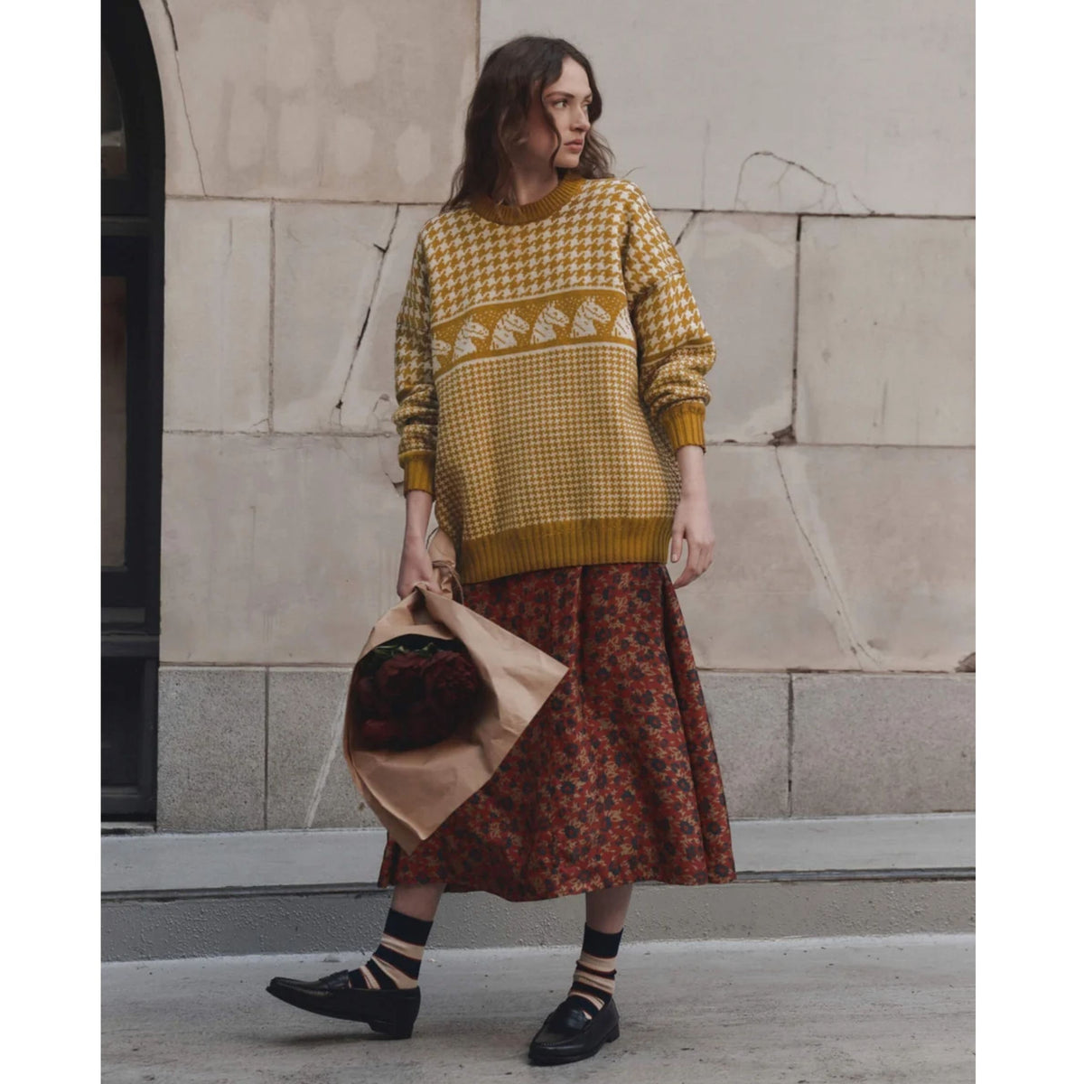 A woman stands outdoors against a stone wall wearing a mustard yellow and white patterned knit sweater, a rust-red floral midi skirt, black loafers, and striped socks, while holding a tan hat.