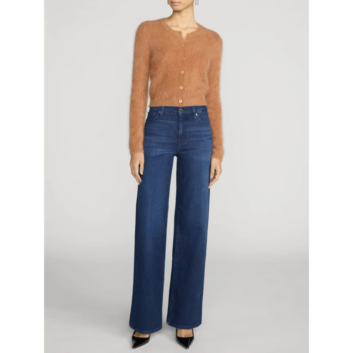 A model wearing a cropped, fuzzy tan cardigan with buttons and high-waisted, wide-leg blue jeans, paired with black pointed-toe heels, set against a plain light gray studio background.