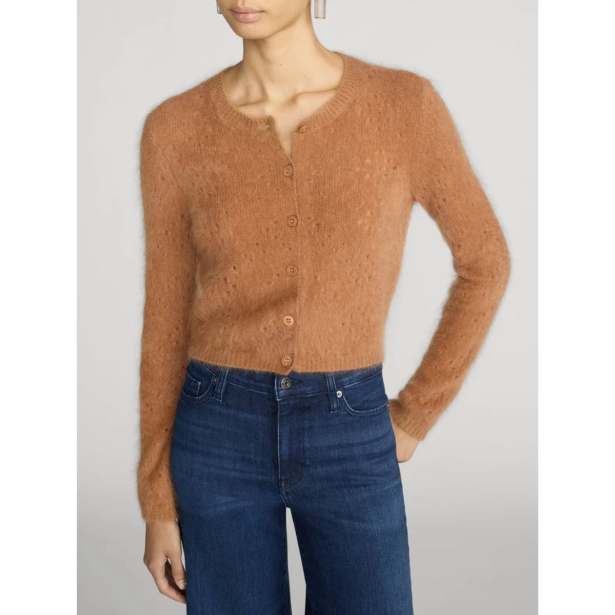 A woman wearing a cropped, fuzzy knit cardigan in a warm camel brown color with matching buttons, paired with dark blue jeans, against a light gray studio background.