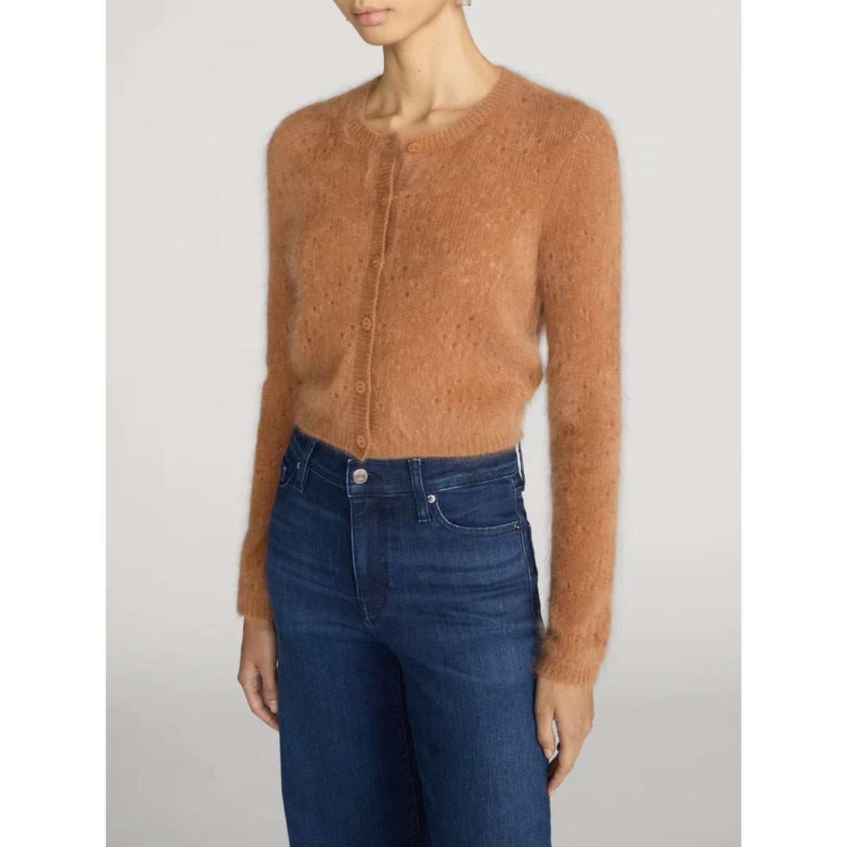 A woman wearing a cropped, fuzzy knit cardigan in a warm camel brown color, paired with dark blue jeans, shown against a light gray studio background.