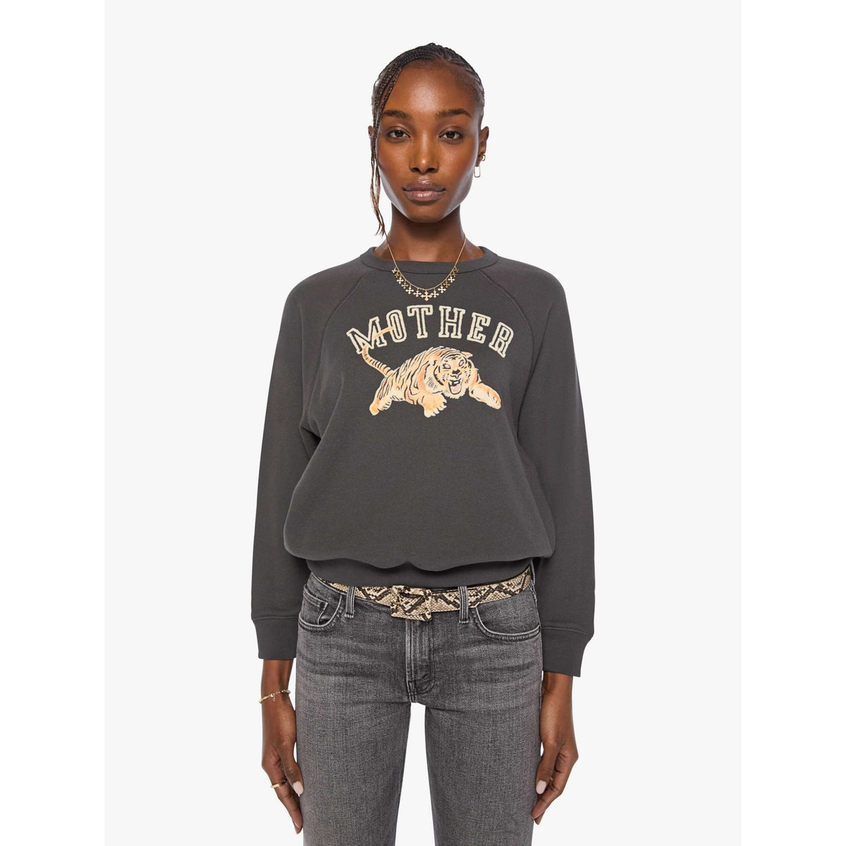 A model wearing a dark gray crewneck sweatshirt with a distressed graphic of a tiger and the word 'MOTHER' in cream, paired with gray jeans and a patterned belt, against a light gray background.