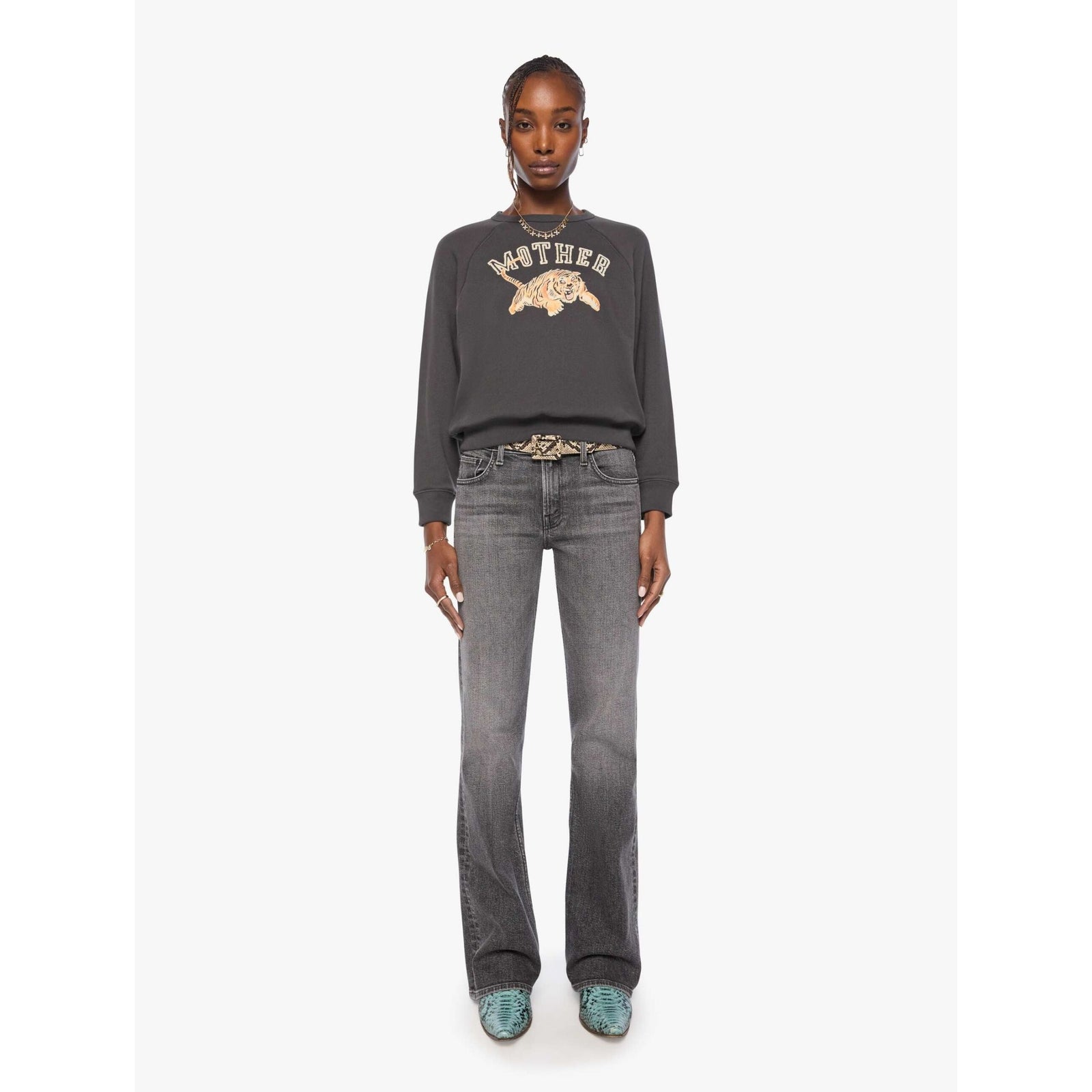 A model wearing a dark gray crewneck sweatshirt with a distressed graphic of a tiger and the word 'MOTHER' in cream, paired with gray jeans and a patterned belt, against a light gray background.