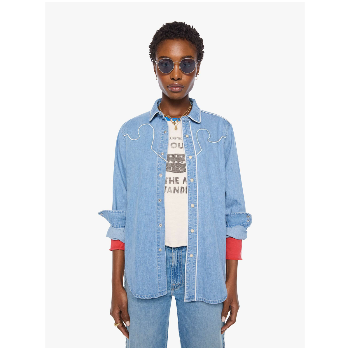 A model wearing a light blue denim shirt with white western-style stitching, layered over a white graphic t-shirt and paired with blue jeans, set against a plain light gray background.