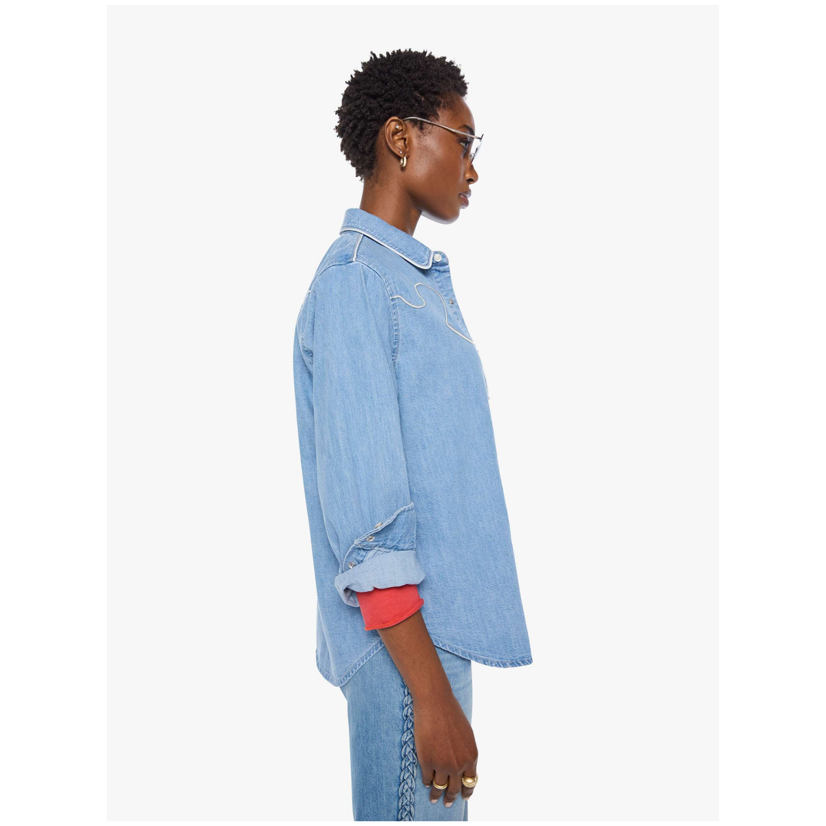 A side-profile view of a model wearing a light blue denim shirt with rolled-up sleeves, showing a bright coral-red inner lining at the cuff, paired with matching blue jeans against a plain white background.