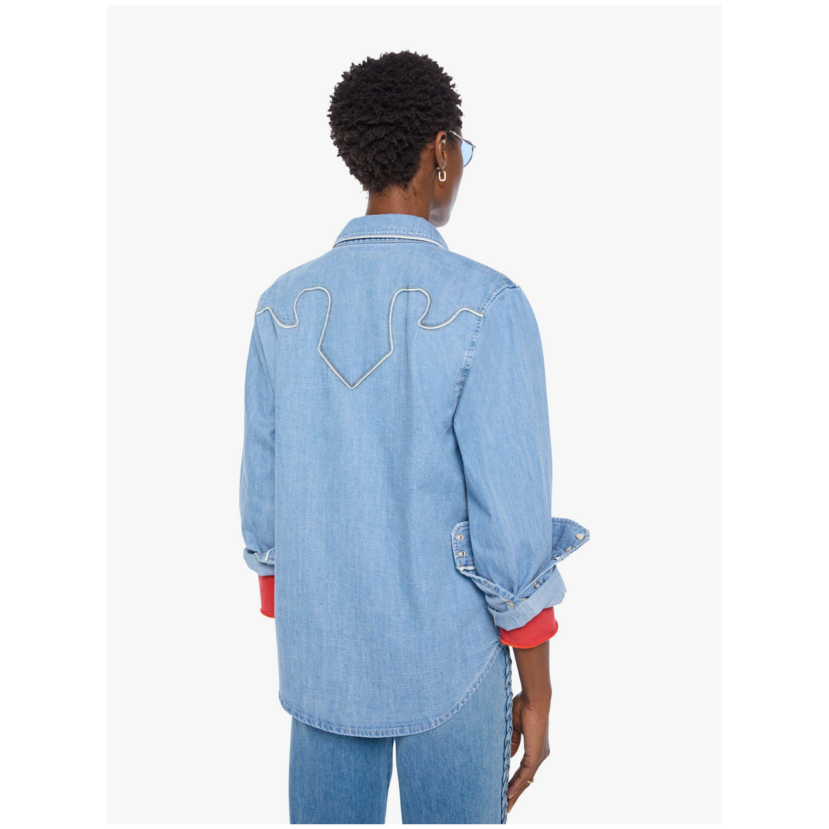 A model shown from the back wearing a light blue denim shirt with a white embroidered design on the upper back, styled with blue jeans and a red undershirt.