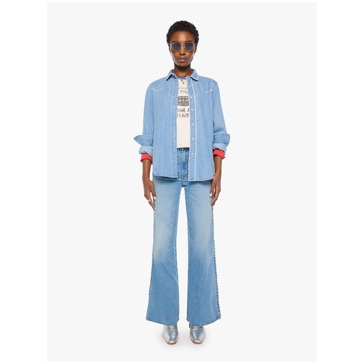 A model wearing a light blue denim set consisting of a button-up shirt jacket and matching wide-leg jeans, styled with a white graphic t-shirt, sunglasses, and silver sneakers against a plain white background.