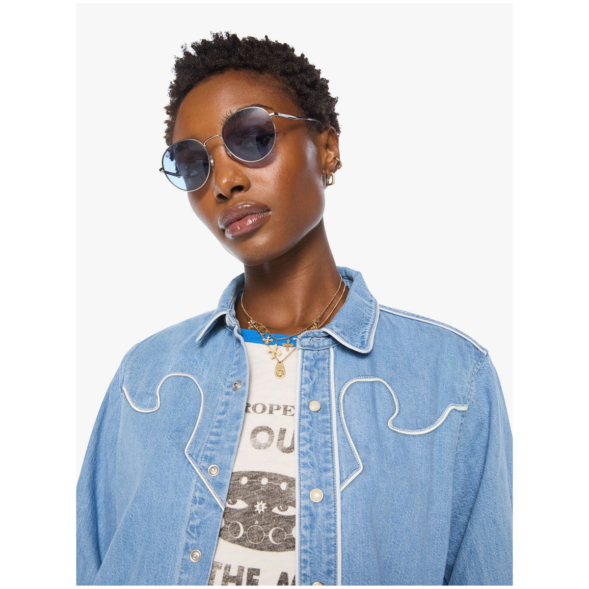 A model wearing round silver-framed sunglasses with dark lenses, a light blue denim shirt jacket with white contrast stitching, and a graphic t-shirt, set against a plain light gray background.