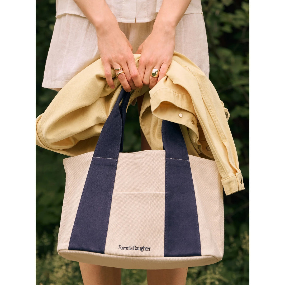 A person outdoors holds a large tote bag with cream canvas sides and navy blue handles and side panels. A light yellow shirt is draped over the bag, and the brand name 'Favorite Daughter' is printed near the bottom front.