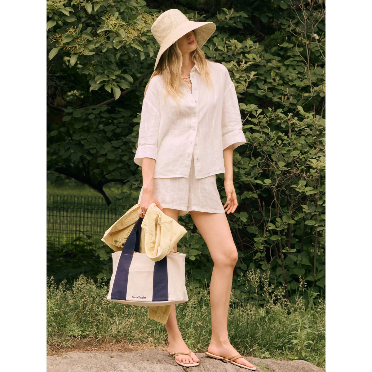 A woman stands outdoors in a garden wearing a light beige wide-brim sun hat, a loose white linen button-up shirt, and matching shorts, holding a navy and cream tote bag.