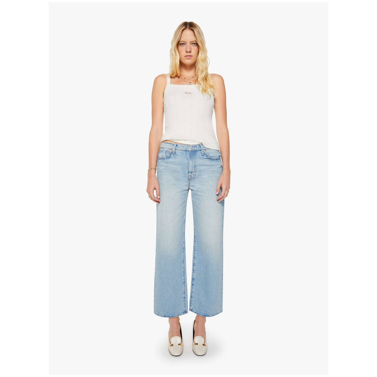 A blonde model stands against a light gray studio background wearing a white sleeveless top, light-wash wide-leg cropped jeans, and white pointed shoes.