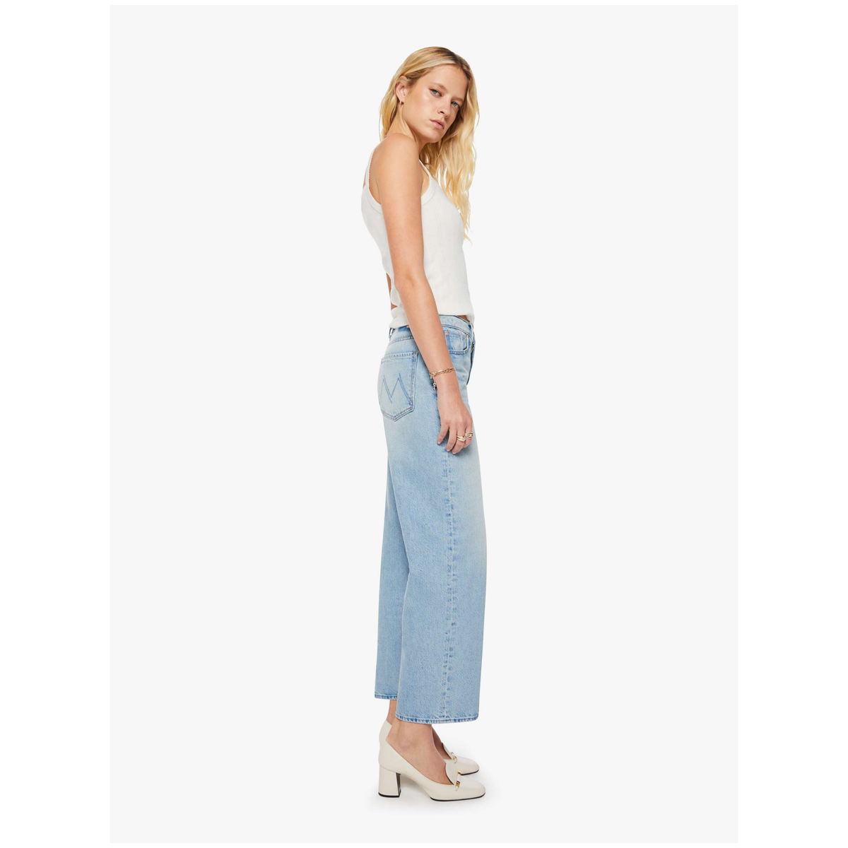 A woman modeling a pair of light-wash wide-leg cropped jeans, paired with a white sleeveless top and white block-heel mules, shown from a side-back angle against a plain white studio background.
