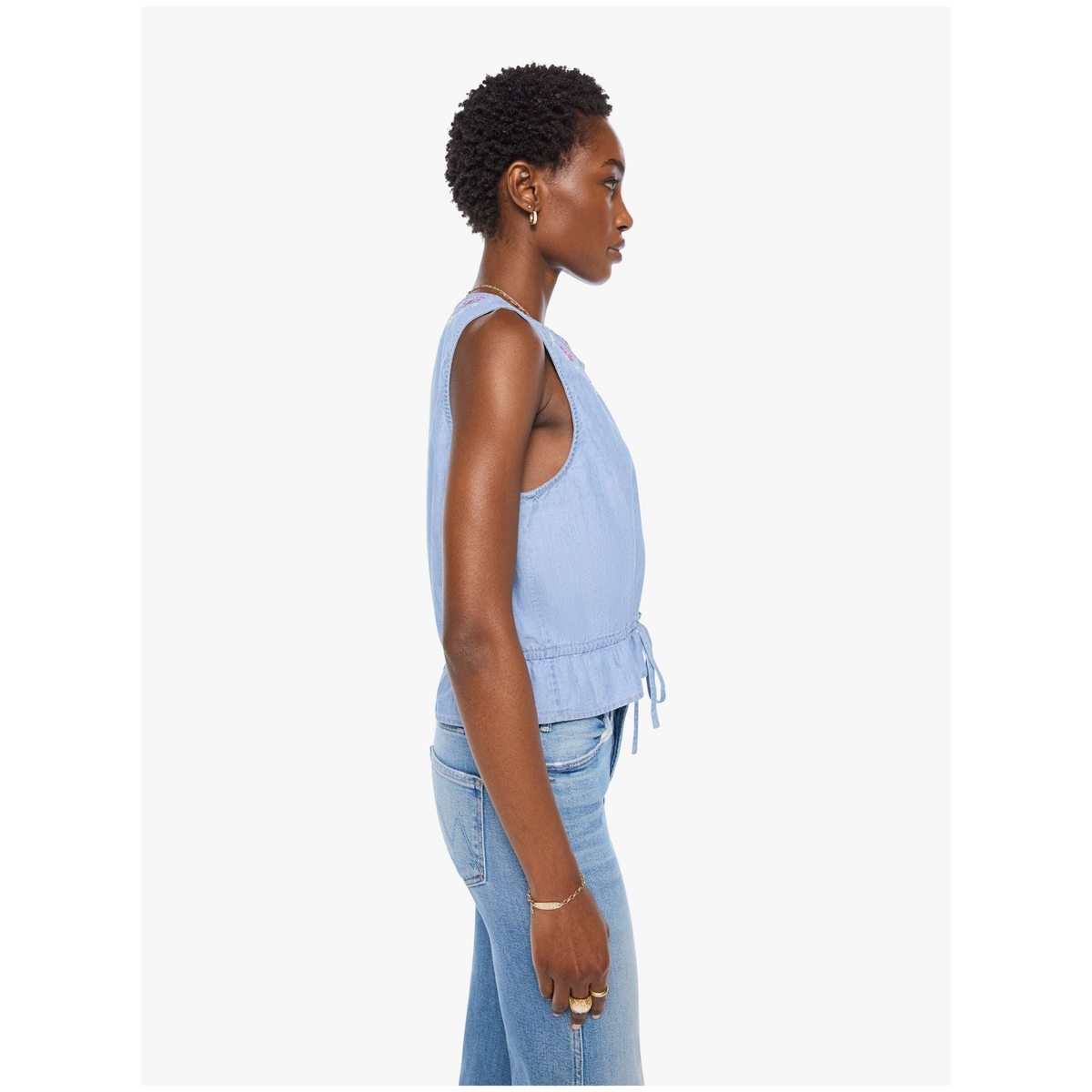 A woman shown in side profile wearing a light blue sleeveless top with a tie detail at the waist and light blue jeans, photographed against a plain light gray studio background.