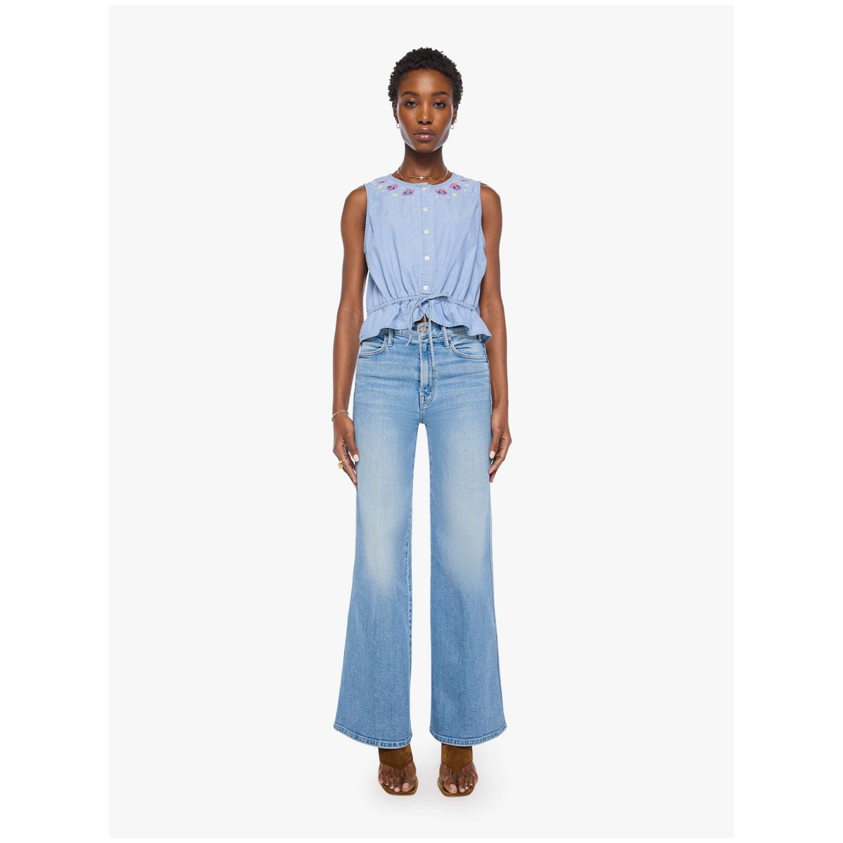 A model stands against a light gray studio background wearing a light blue sleeveless top with a button front and tie detail, paired with light blue wide-leg denim jeans and brown heeled sandals.