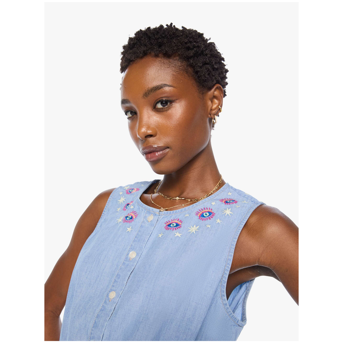 A woman wearing a light blue sleeveless button-front top with colorful embroidered eye motifs around the neckline, photographed against a plain light gray studio background.
