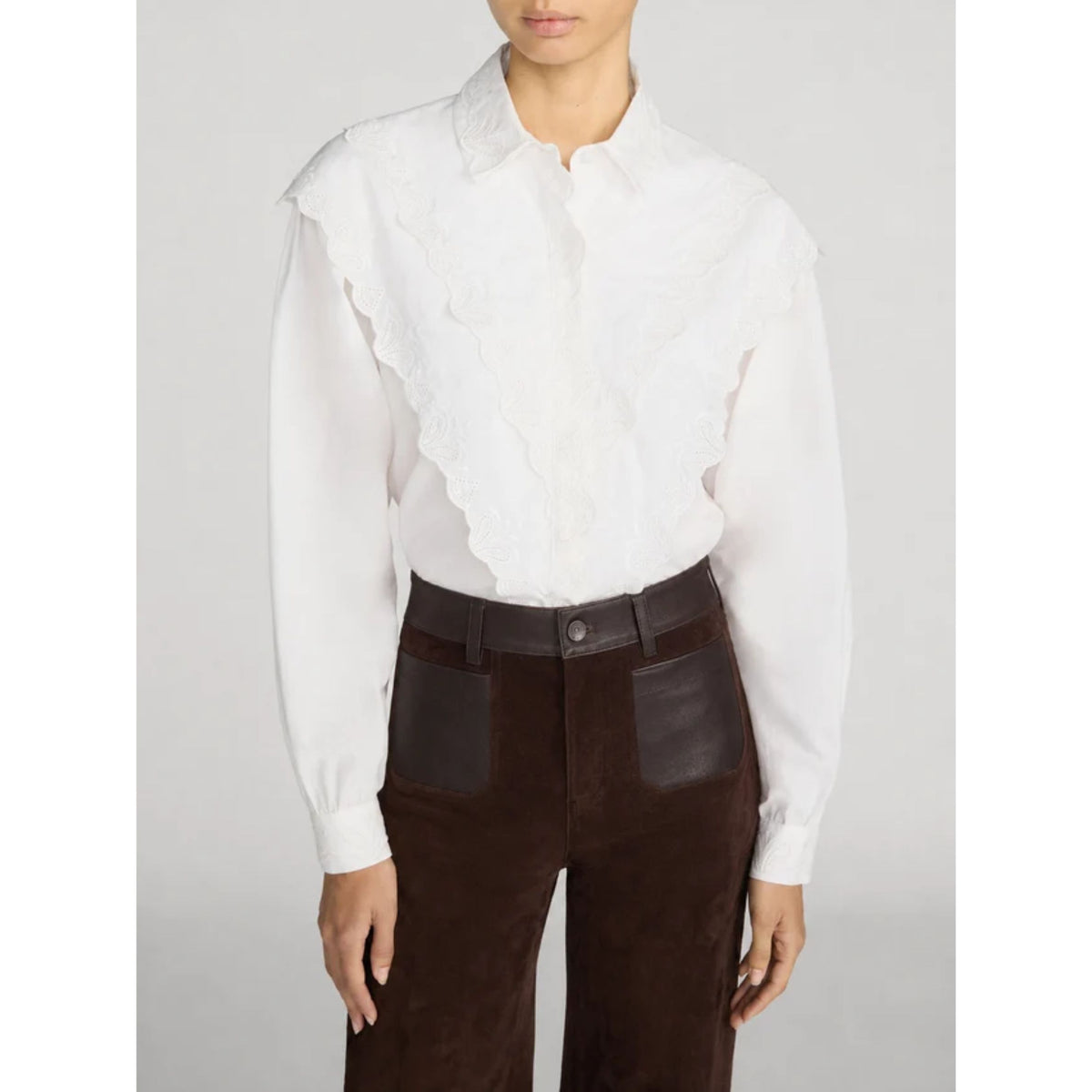 A model wearing a white long-sleeve blouse with a ruffled lace-like front panel and dark brown high-waisted trousers with a dark brown belt.