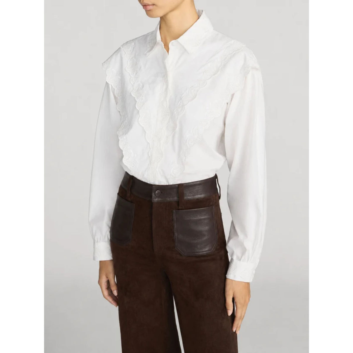 A model wearing a white long-sleeve blouse with ruffled lace detailing on the front and dark brown corduroy trousers with a dark brown belt.