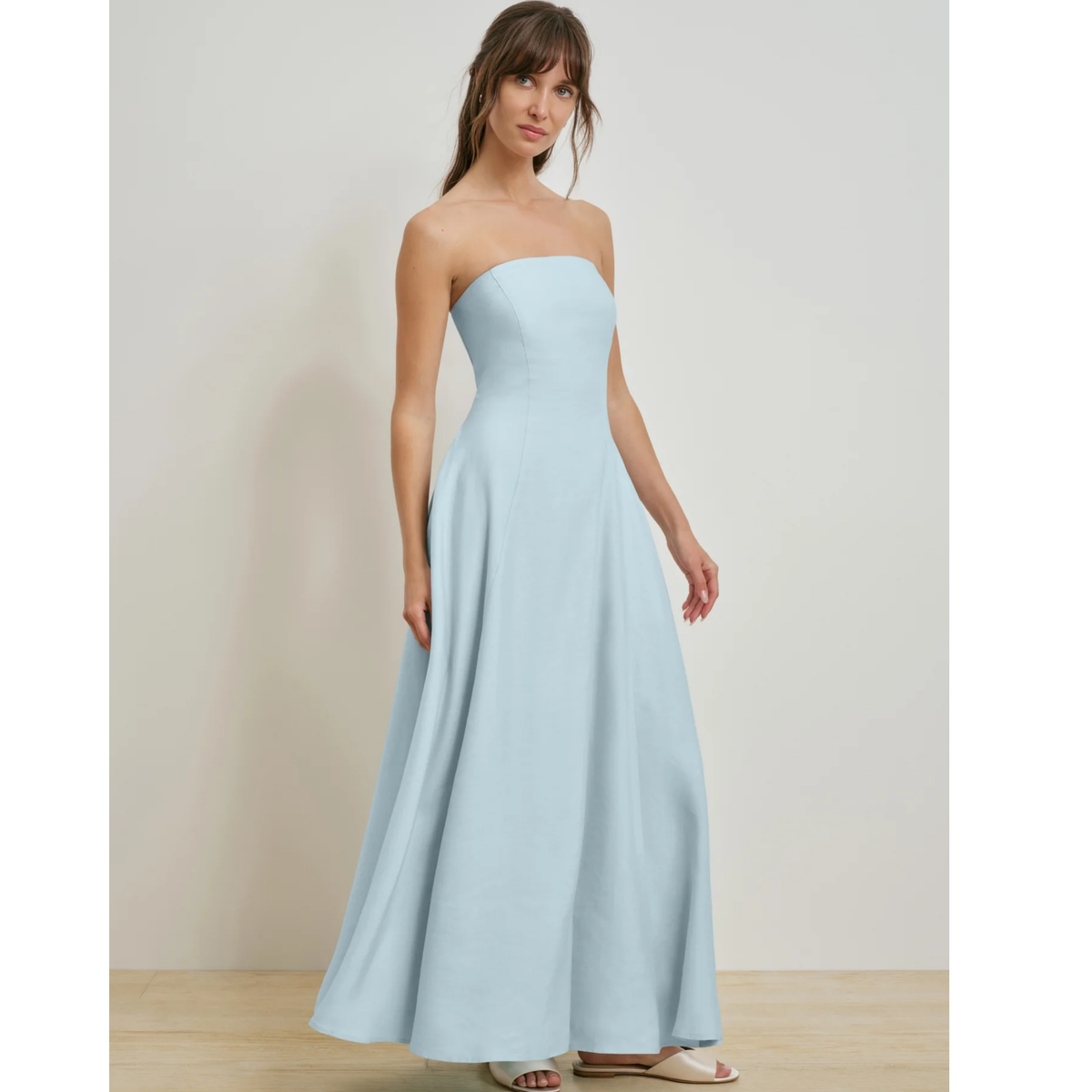 A woman modeling a strapless light blue maxi dress with a fitted bodice and flowing skirt, standing against a plain light beige wall.