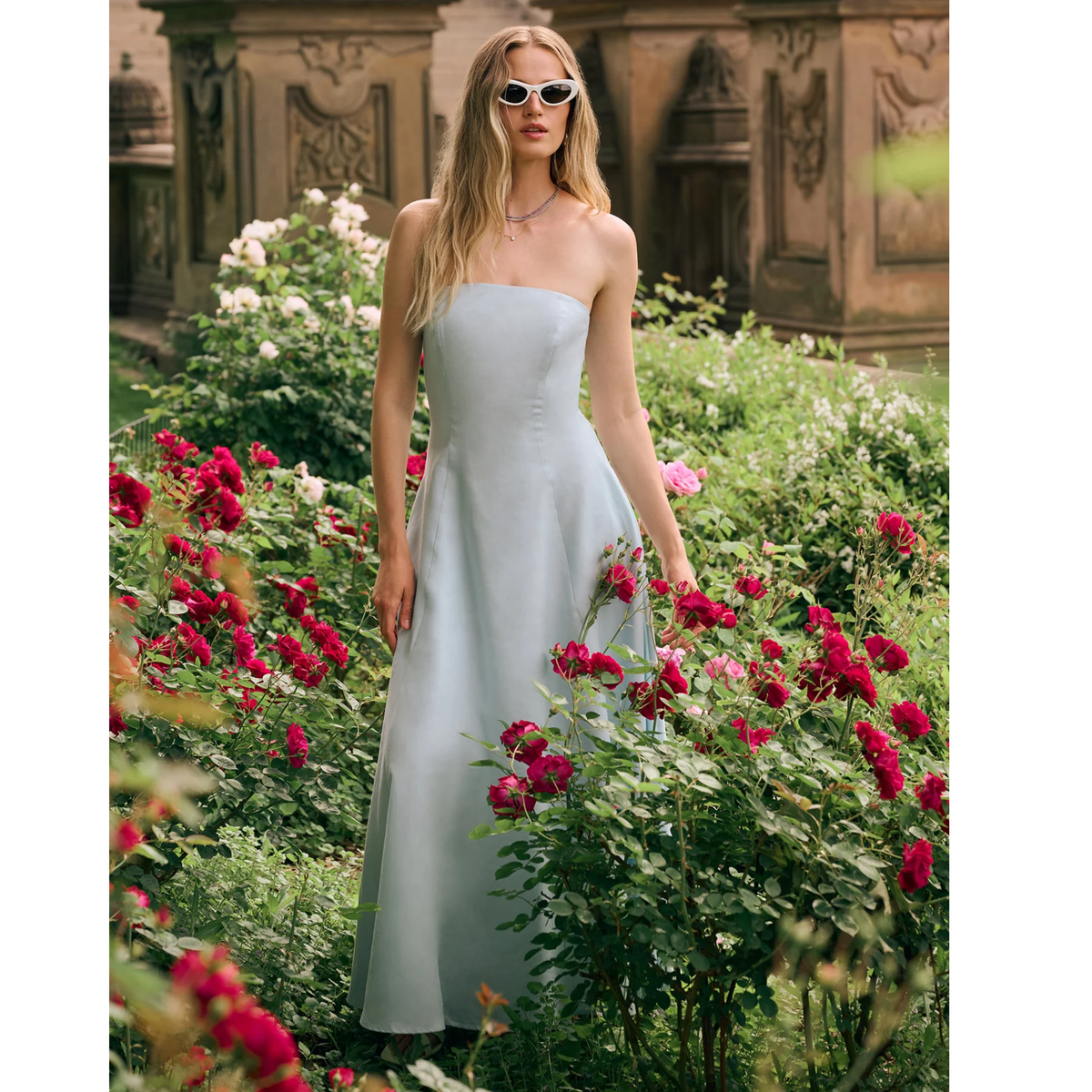 A woman wearing a strapless light blue midi dress and white-framed sunglasses stands in a garden surrounded by red and white flowers, with a stone building in the background.