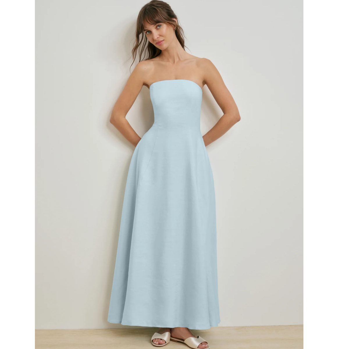A woman modeling a strapless light blue maxi dress with a fitted bodice and full skirt, standing against a plain light beige wall.