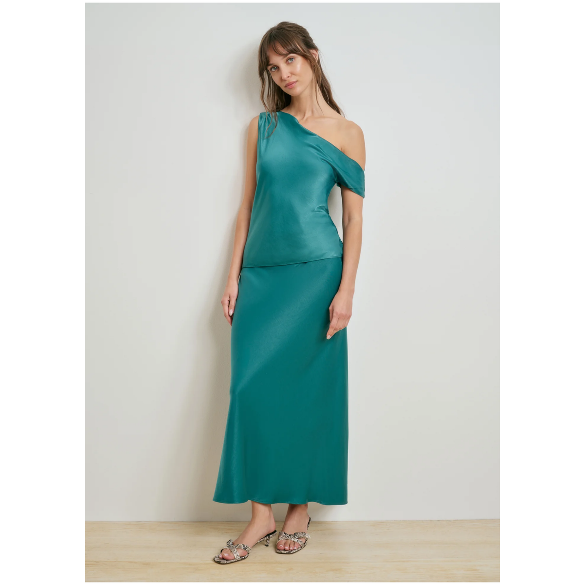 A woman modeling a teal green one-shoulder satin midi dress with a matching skirt, standing against a neutral light gray wall.