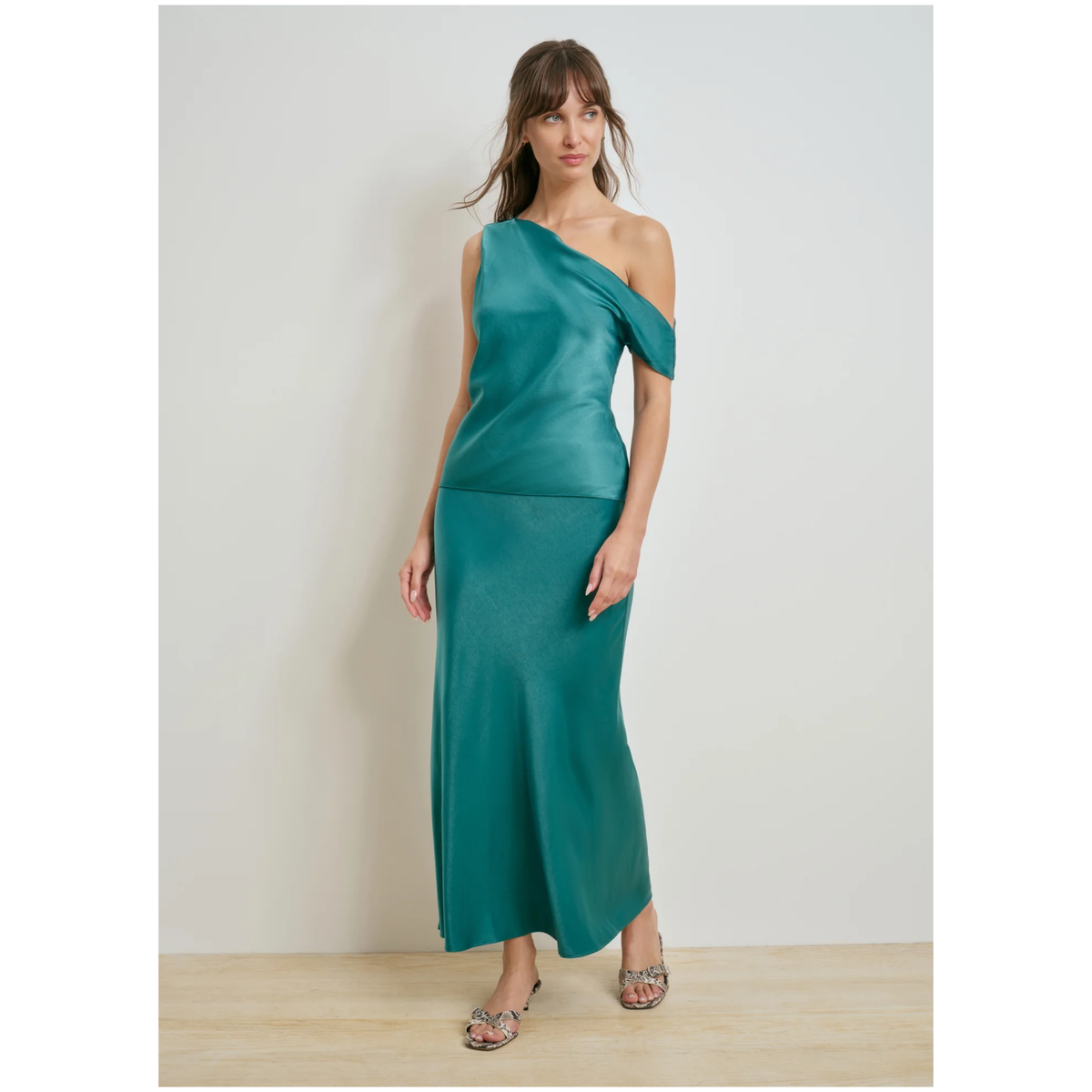 A woman modeling a teal green one-shoulder satin maxi dress with a draped neckline and a flowing skirt, standing against a neutral light gray wall and light wood floor.