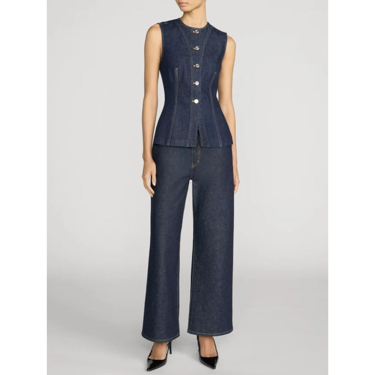 A model wearing a dark indigo denim two-piece set consisting of a sleeveless button-front vest and matching wide-leg pants, paired with black pointed-toe heels, against a light gray studio background.