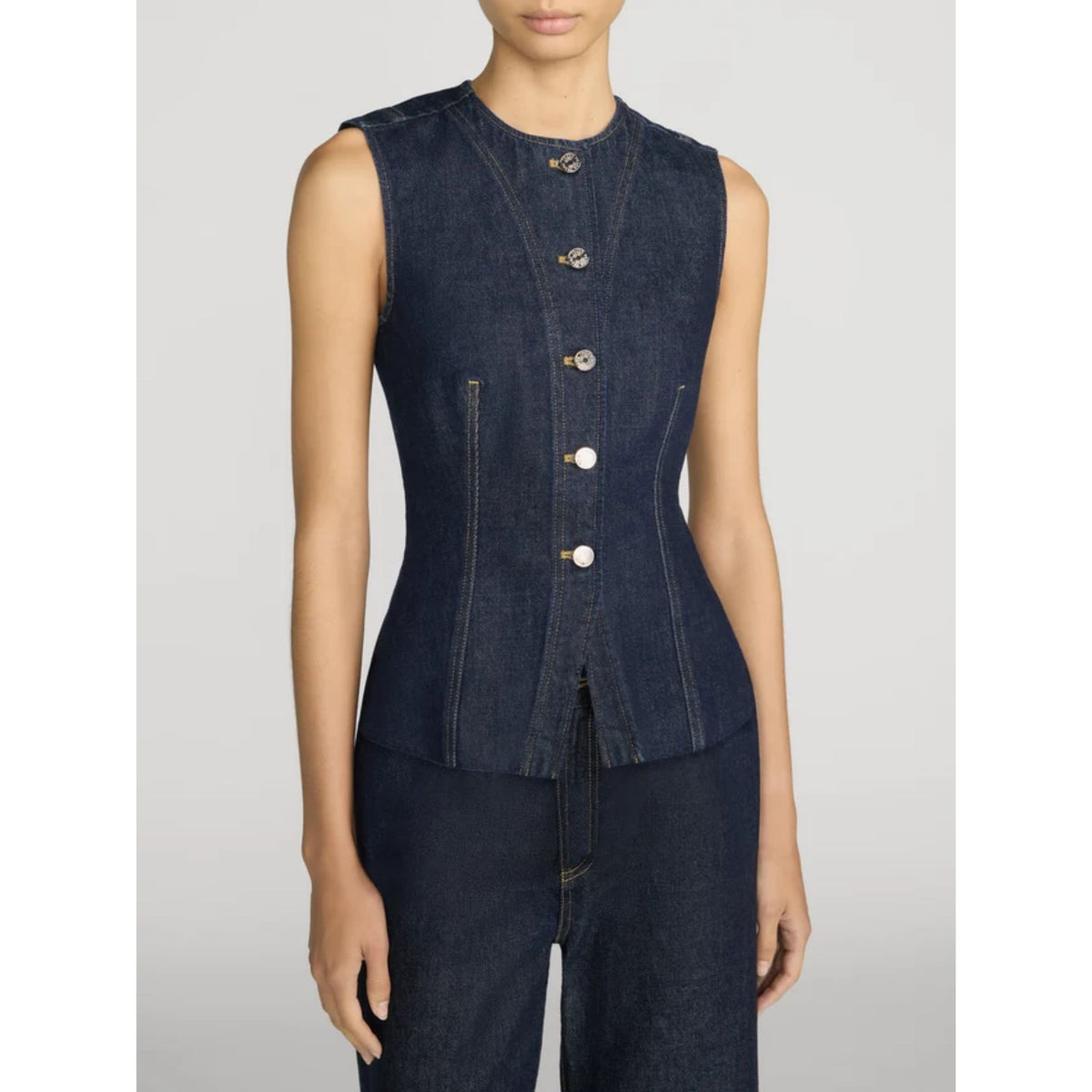 A model wearing a sleeveless dark indigo denim vest with decorative buttons down the front, paired with matching dark denim pants, against a light gray studio background.
