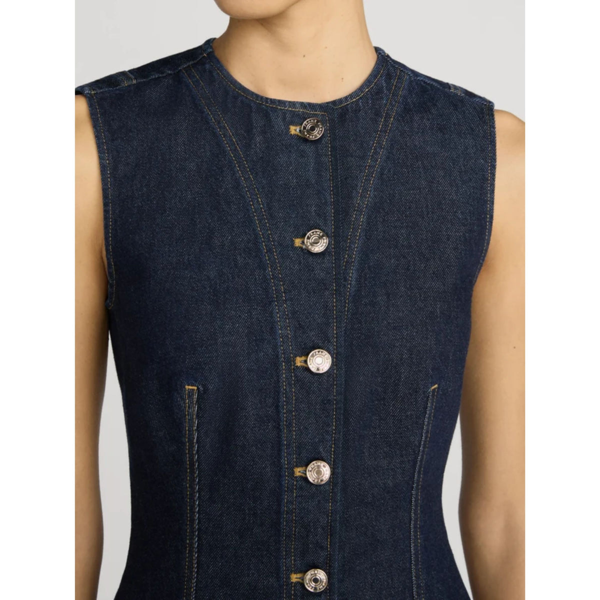 Close-up of a sleeveless dark indigo denim vest with a high round neckline, front button closure, and zippered side pockets, shown on a model against a light gray background.