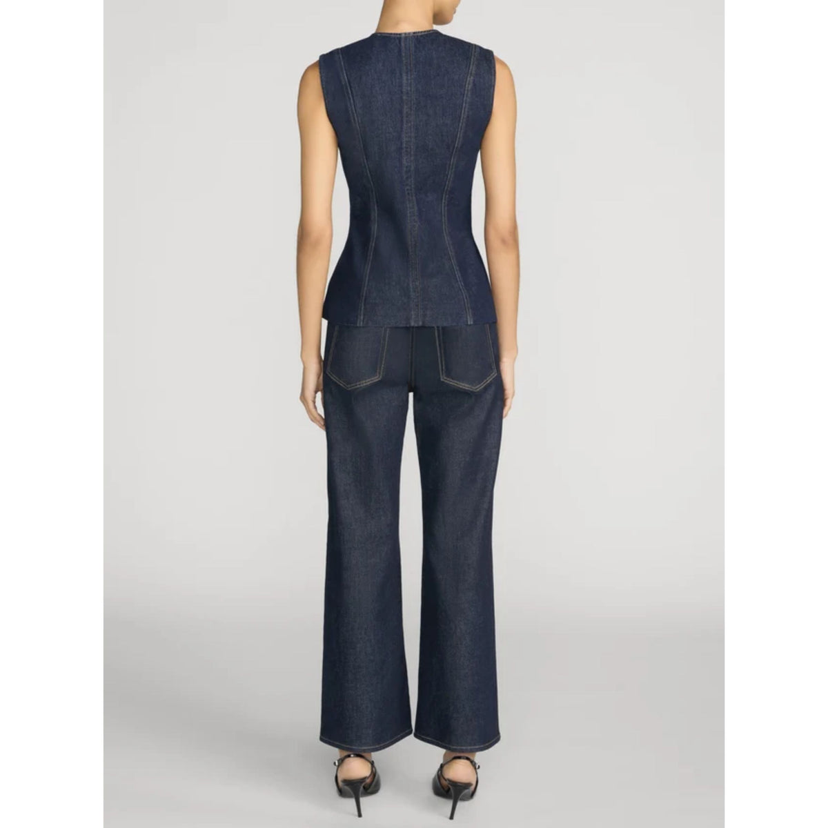 A model shown from the back wearing a dark indigo denim sleeveless peplum top and matching wide-leg denim pants, paired with black high heels, against a light gray studio background.