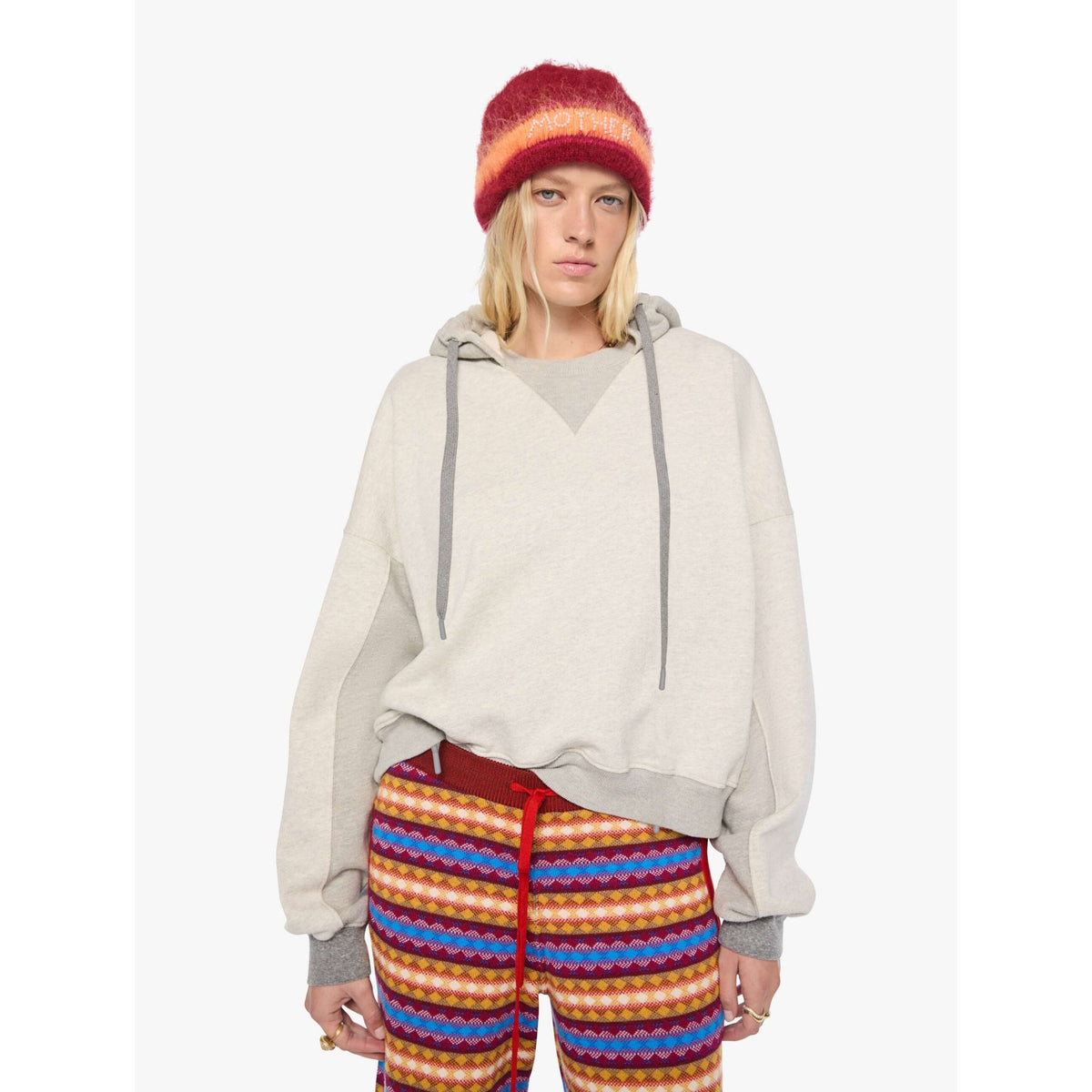 A model wearing a light beige oversized pullover hoodie with gray drawstrings, a red and orange striped knit beanie, and colorful striped pants against a plain white background.