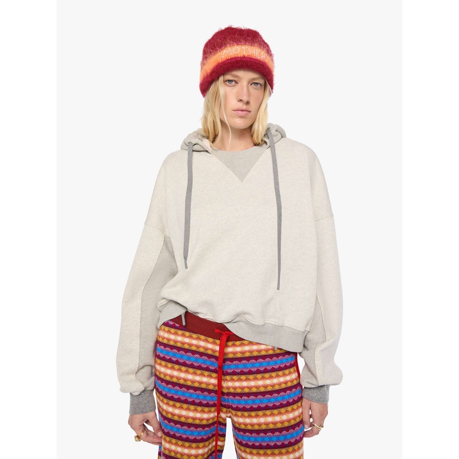 A model wearing a light beige oversized pullover hoodie with gray drawstrings, a red and orange striped knit beanie, and colorful striped pants against a plain white background.