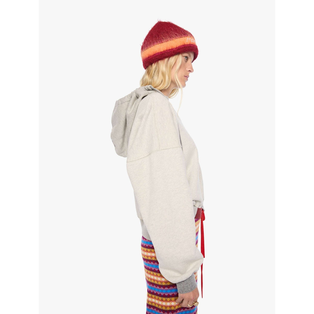 A model shown from the side profile wearing a light gray oversized hoodie, a red and orange striped knit beanie, and colorful striped pants against a plain white background.