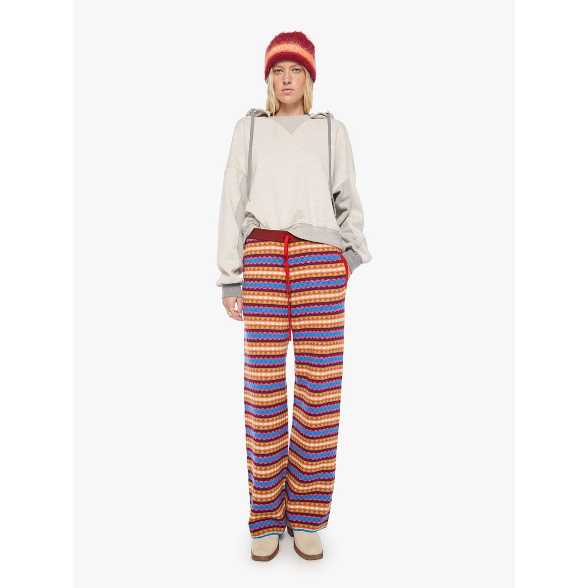 A model stands against a light gray studio background wearing a light gray hoodie, a red knit beanie, and wide-leg knit pants with horizontal stripes in red, blue, orange, and yellow.