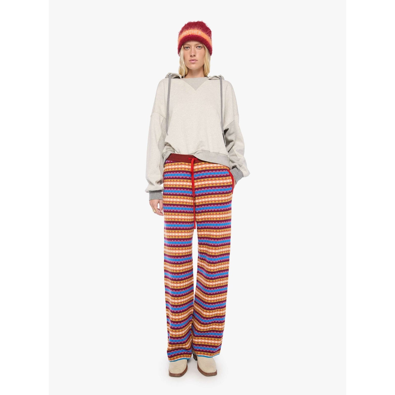 A model wearing a light beige oversized pullover hoodie with gray drawstrings, a red and orange striped knit beanie, and colorful striped pants against a plain white background.