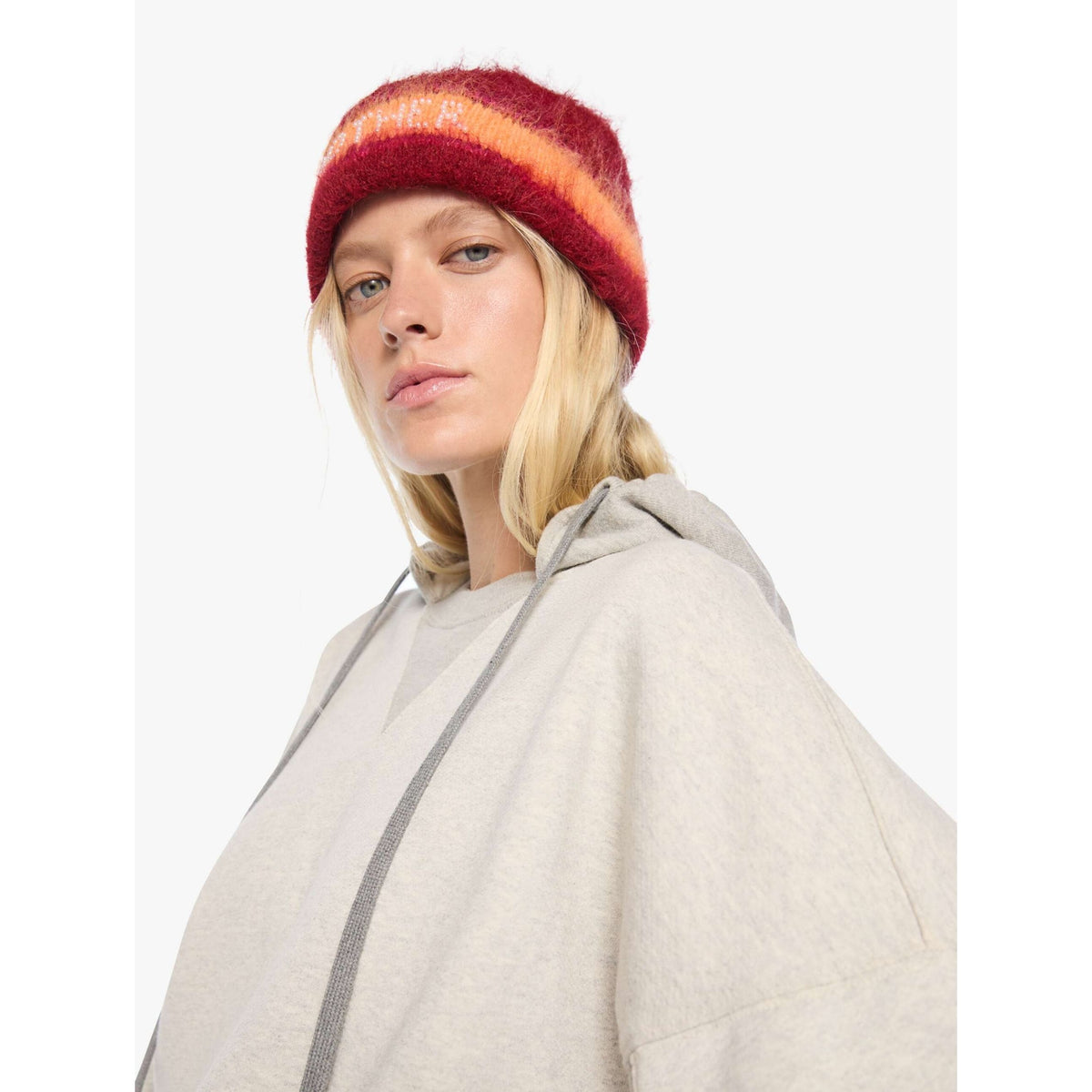 A woman wearing a fuzzy knit beanie with horizontal stripes in red, orange, and dark red, paired with a light gray hoodie, photographed against a plain light gray background.