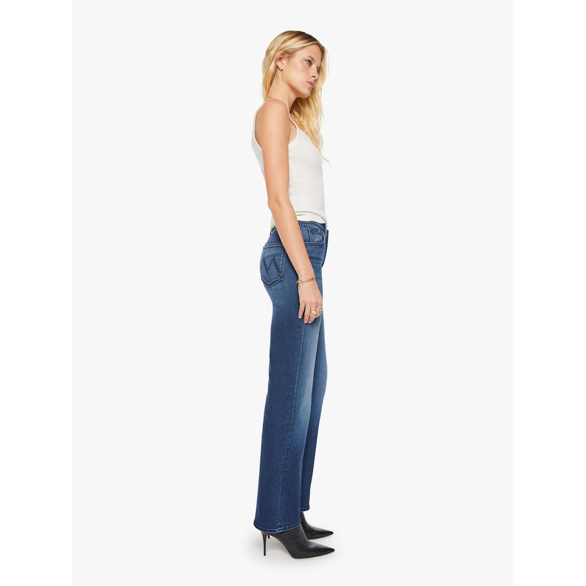 A woman shown in side profile wearing blue wide-leg jeans, a white sleeveless top, and black pointed-toe heels against a plain light gray studio background.