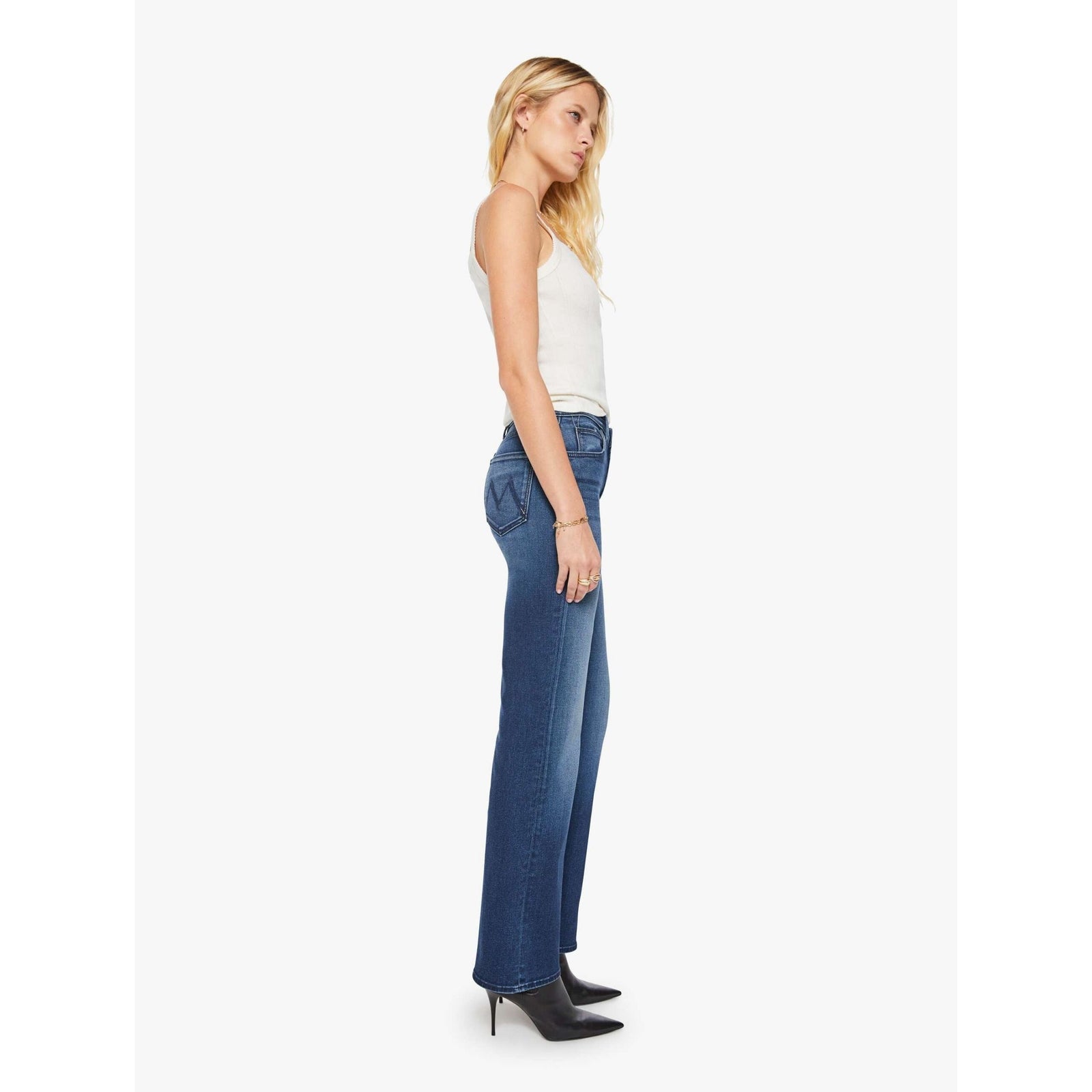 A woman modeling a pair of medium-wash blue jeans with a straight-to-slightly-flared leg, paired with a white tank top and black ankle boots, against a plain white studio background.