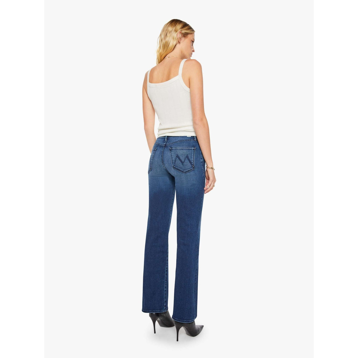 A woman shown from the back wearing blue flare jeans, a white ribbed tank top, and black pointed-toe heels against a plain light gray studio background.