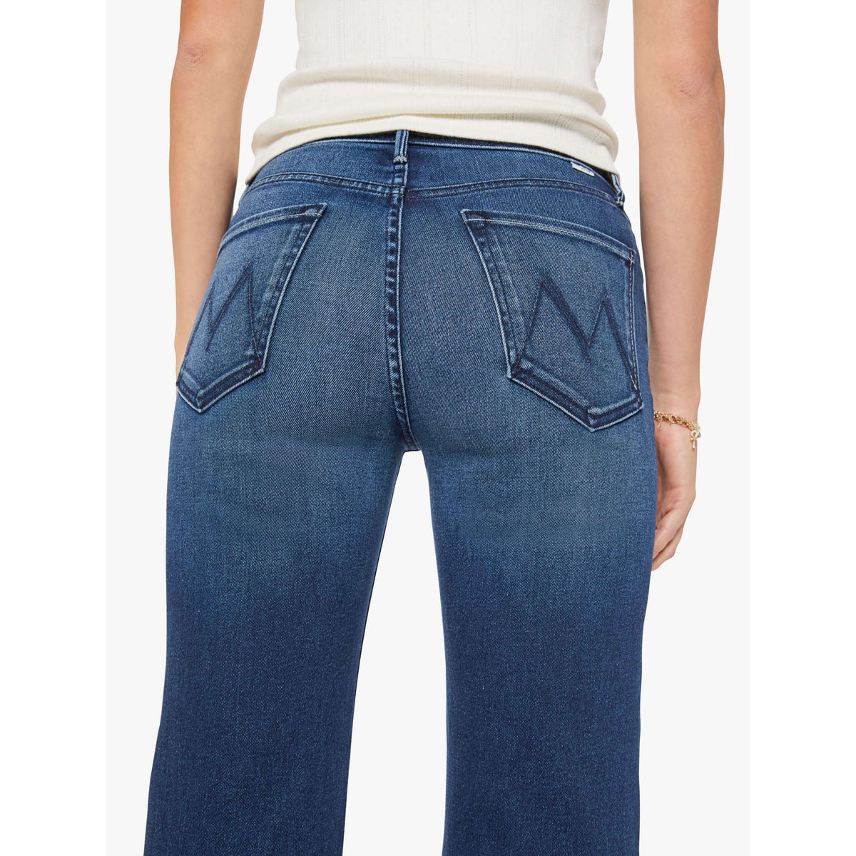 Rear view of a person wearing blue denim jeans with a large embroidered 'M' on each back pocket, paired with a cream ribbed top against a light gray studio background.