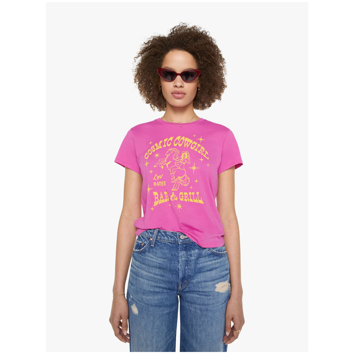 A woman modeling a bright pink graphic t-shirt with yellow text that reads 'Cocaine Cowboys' and 'Bad & Gell', paired with distressed blue jeans against a light gray background.