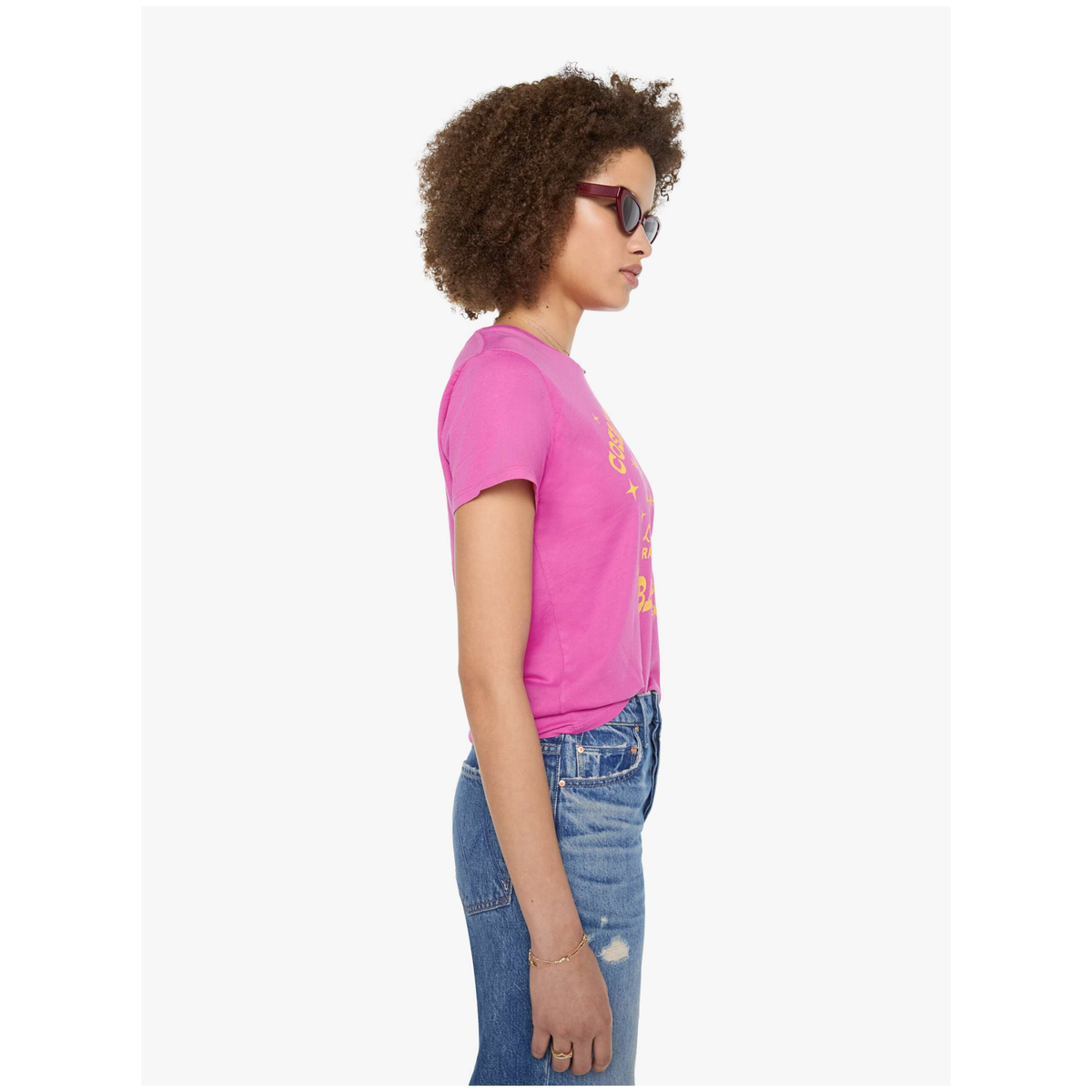 A woman in profile wearing a bright pink short-sleeve t-shirt with yellow graphic text and blue jeans, photographed against a plain light gray background.