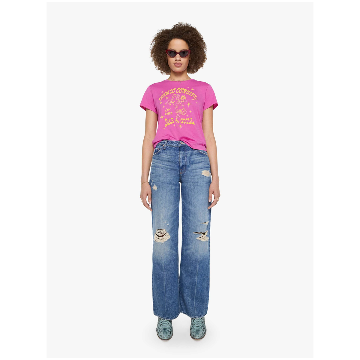 A model wearing a bright pink graphic t-shirt, blue wide-leg distressed jeans, and teal sneakers, standing against a plain light gray background.