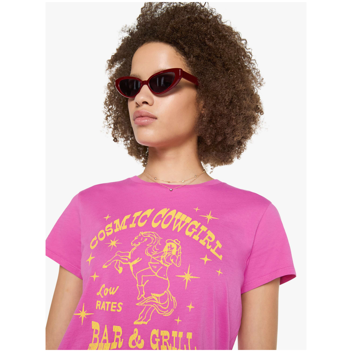 A model with curly brown hair wearing a bright pink graphic t-shirt with yellow text reading 'COSMIC COWGIRL' and 'LOW RATES BAR & GRILL', along with a line drawing of a cowgirl on a horse. She is also wearing dark red cat-eye sunglasses against a plain white background.