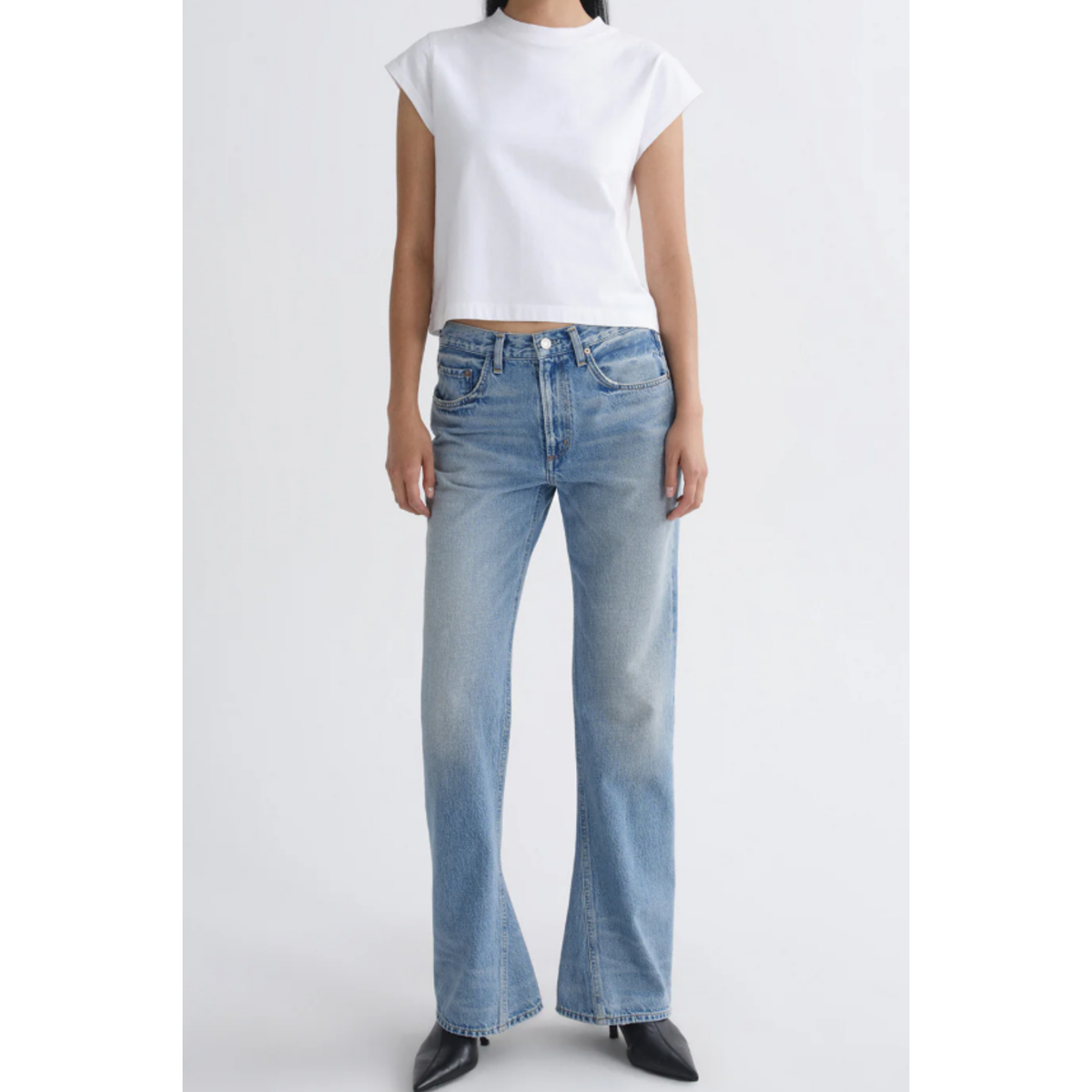 A model wearing a white cropped short-sleeve t-shirt and light-wash blue flare jeans with black pointed-toe heels, shown against a plain light gray studio background.