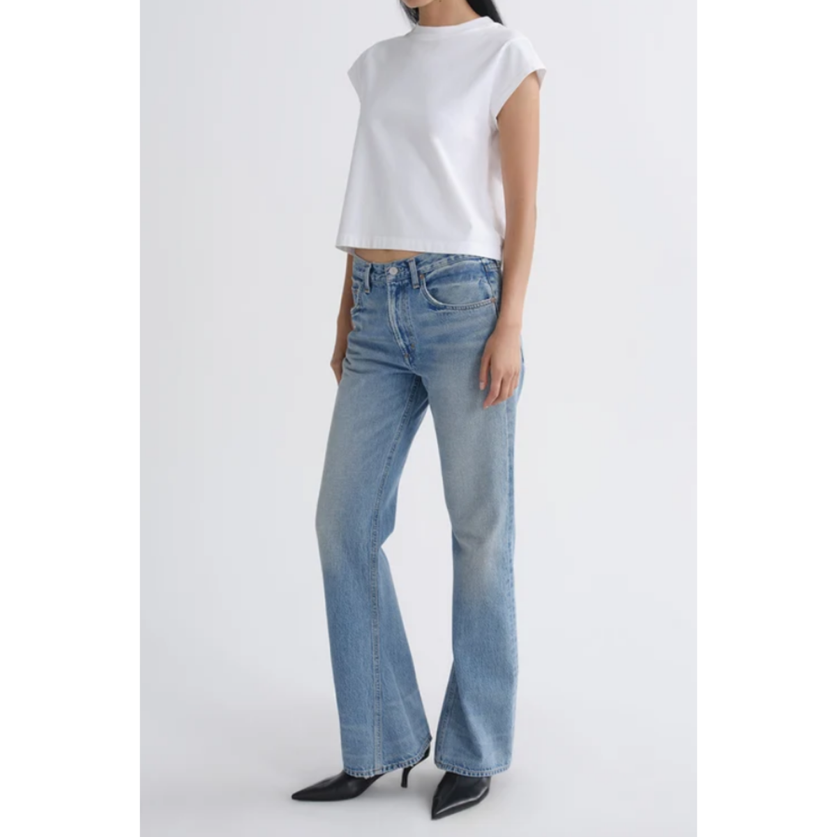 A model wearing light-wash blue flare jeans with a white cropped short-sleeve top and black pointed-toe heels, standing against a light gray studio background.