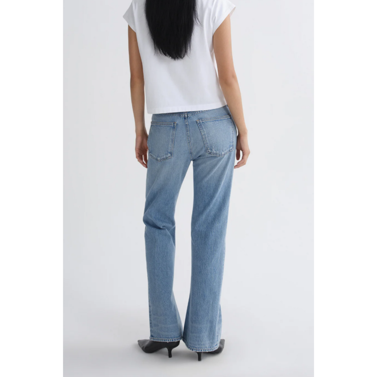 A model shown from the back wearing light-wash blue flare jeans, a white t-shirt, and black heels against a plain light gray studio background.