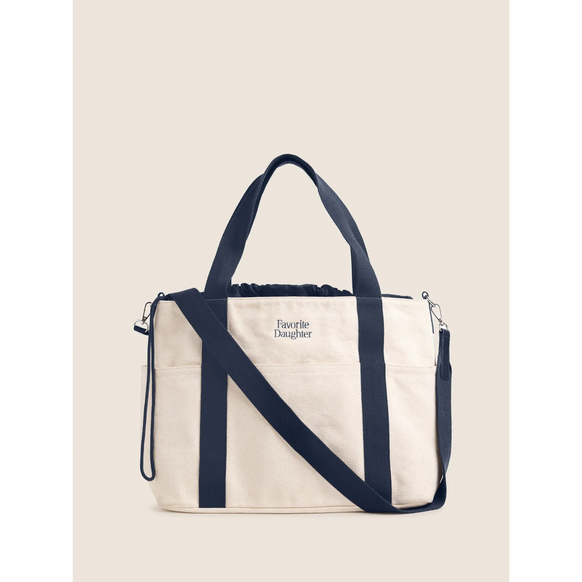 A cream canvas tote bag with navy blue straps and trim, featuring the text 'Favorite Daughter' on the front, set against a solid beige background.