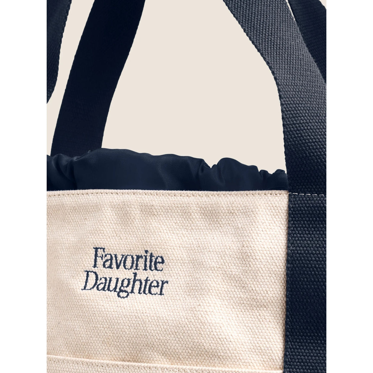 Close-up of a canvas tote bag with dark navy blue straps and trim, featuring an off-white woven body with the text 'Favorite Daughter' printed in navy on the front.