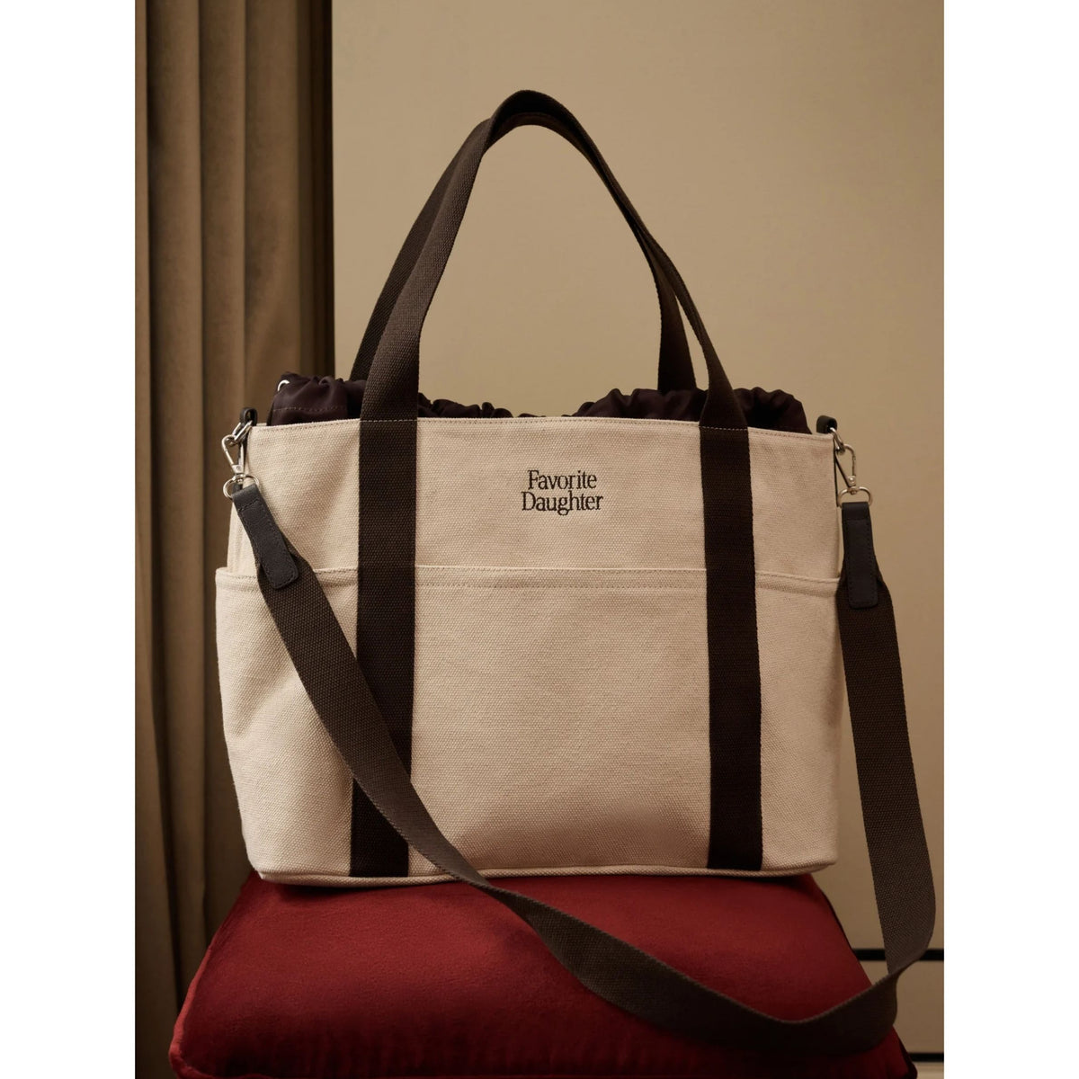 A canvas tote bag in beige with dark brown straps and trim, featuring the text 'Favorite Daughter' on the front. The bag is displayed on a red velvet cushion against a neutral beige background.