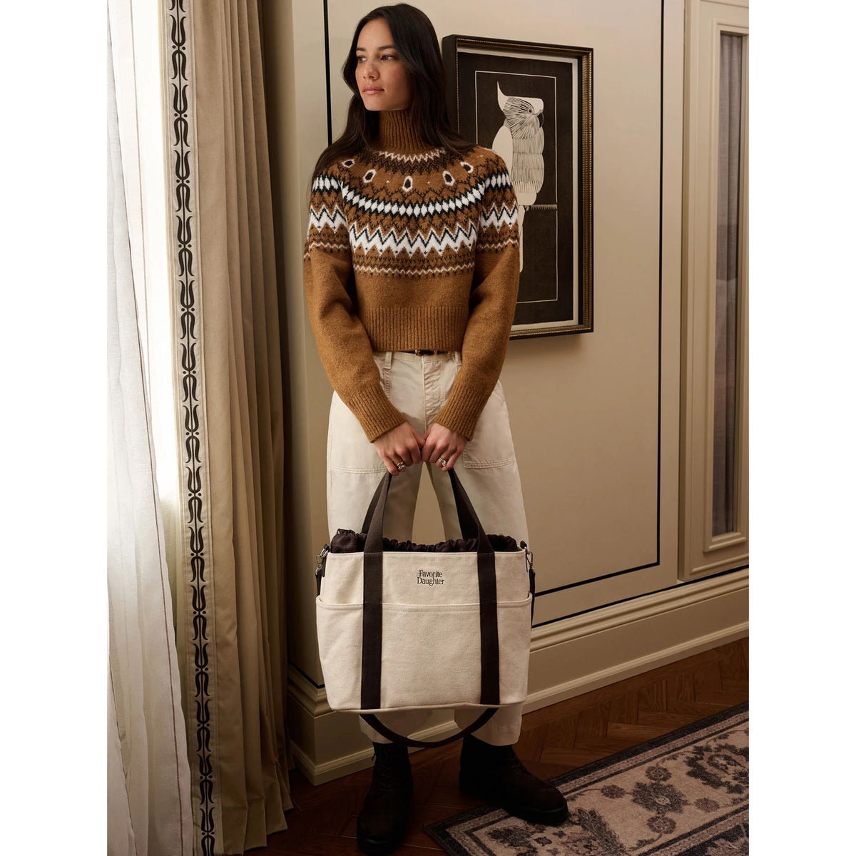 A woman stands indoors holding a large cream canvas tote bag with dark brown handles and trim. She wears a brown Fair Isle knit sweater, off-white pants, and dark boots.